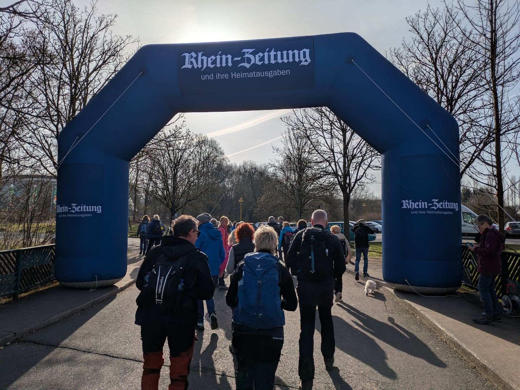 A group of people with backpacks walk under a large blue inflatable archway labeled “Rhein-Zeitung” on a sunny day, with bare trees and a clear sky in the background.